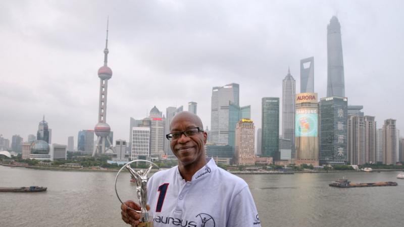 A man's upper body in front of a skyline with skyscrapers in the background. He is holding a silver statue to the camera, smiling