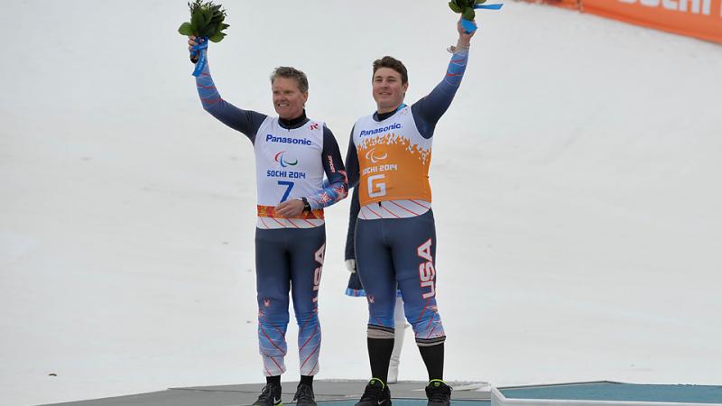 Two men in skiing suits waving flower bouquets. They both stand on a podium in the snow