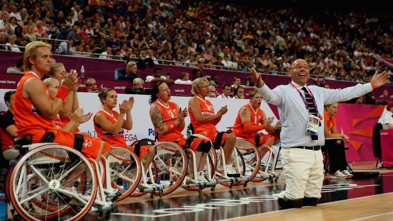 Female wheelchair basketball players lined up, cheering during a game with an amputee man in a suit standing in front of them