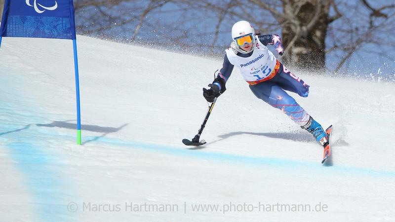 Skier with one leg skis at speed past a gate with her outrigger outstretched