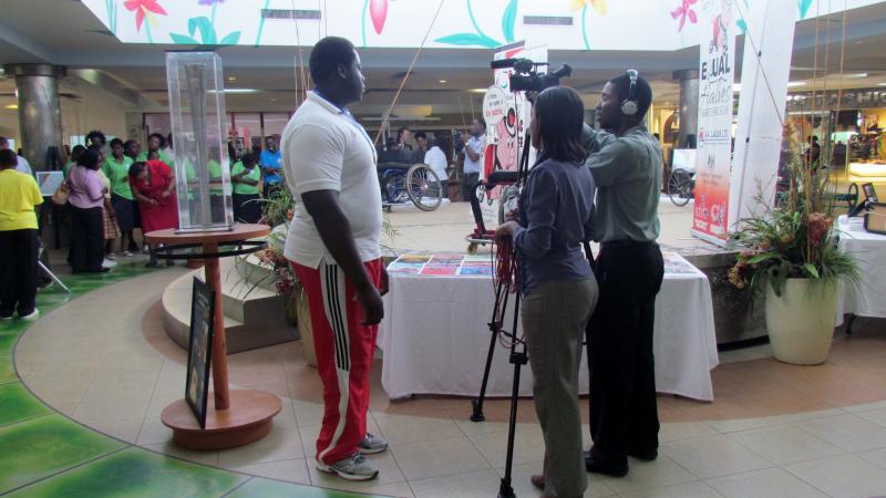 Team of a camera man and a women holding a microphone stand opposite of a man in sport dress. They seem to be preparing an interview. Background: A stage with a wheelchair on it