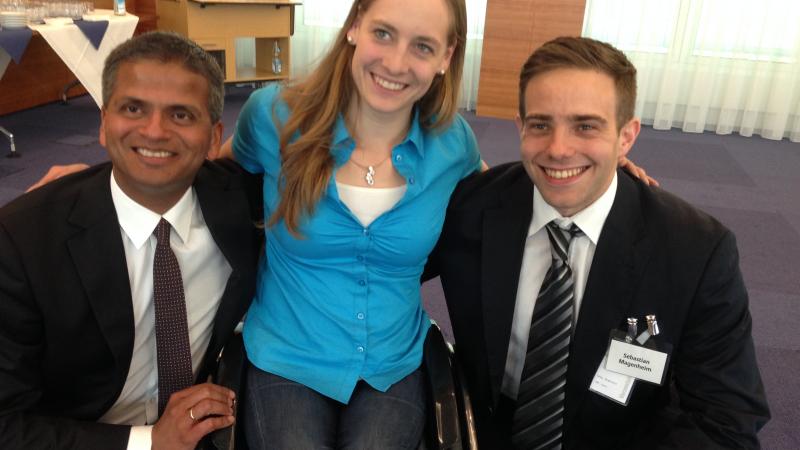 Women in wheelchair smiling, poses surrounded by two men in suits who perch next to he