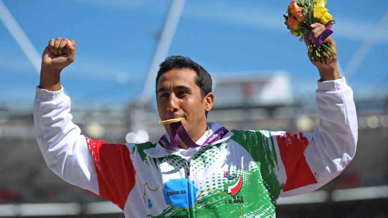 Man in training suit biting on a medal, waving arms in the air. He is holding a flower bouquet and is in a stadium.