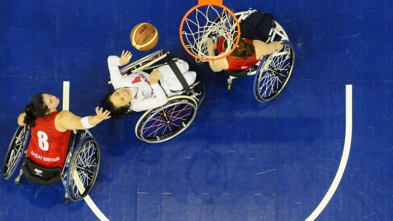 Picture taken from above the basketball basket. Three female players in wheelchairs fighting for the ball under the basket.