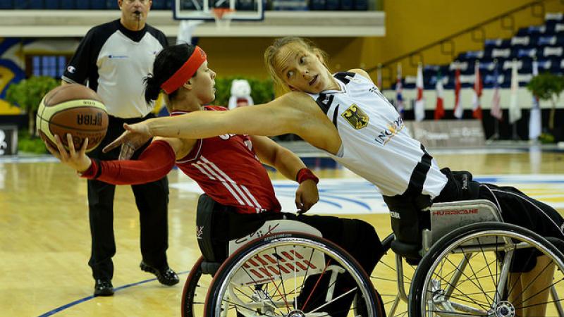 Two female basketball players in wheelchairs fight for the ball. A referee watches them from behind
