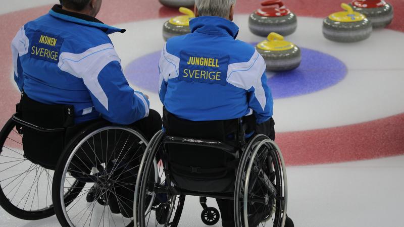 Two men in wheelchair on ice, looking at some curling stones.