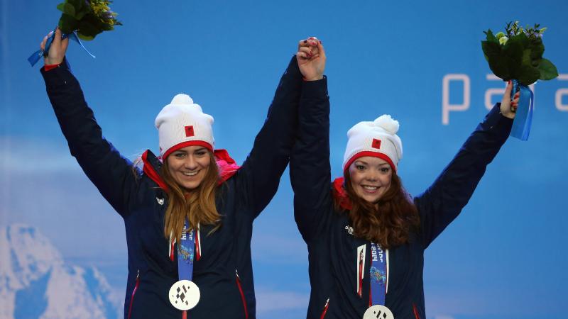 Two young women wearing ski clothes and a silver medal holding hands and waving