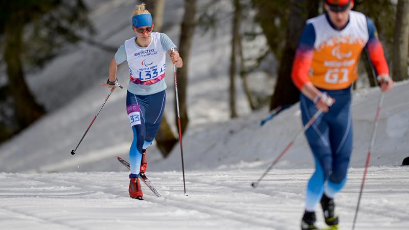 Female cross country skier behind a male cross country skier with orange bib on a track