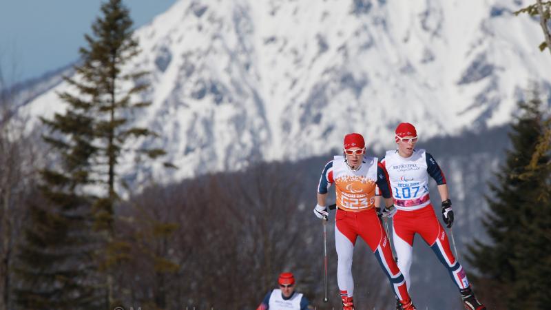 Wide shot of two Nordic skiers skating close together on the crest of the hill, with one skier a short distance behind. Big mountain in background