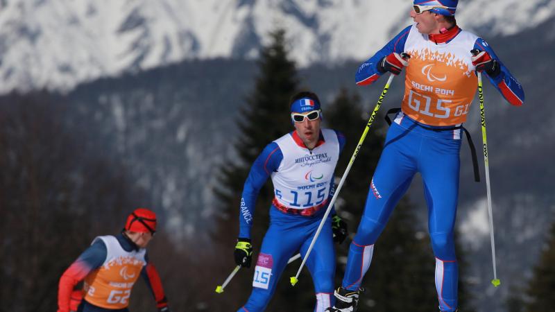 A Nordic skier looks behind him at two other skiers approaching the crest of the hill, close behind him. Mountains are in the background.