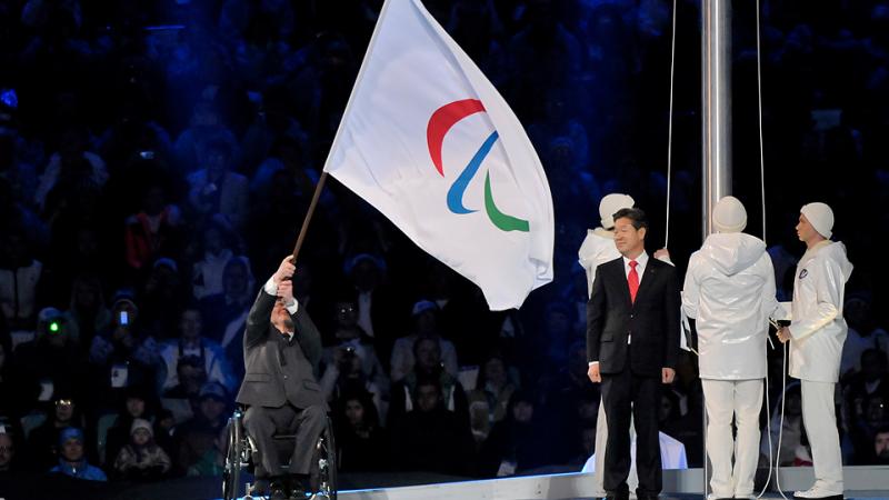 Man in wheelchair on a stage waving a huge flag with the Paralympic symbol on it, handing it over to another person.