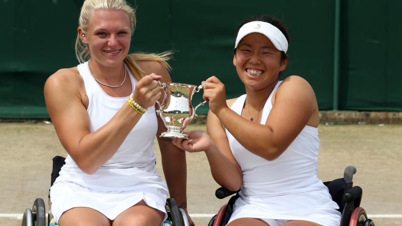 Two women in wheelchairs on a tennis court hold a trophy and smile to the camera.