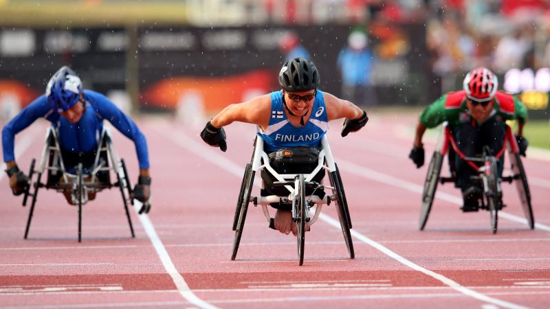 Three wheelchair racers approaching the finish line on the track of a stadium