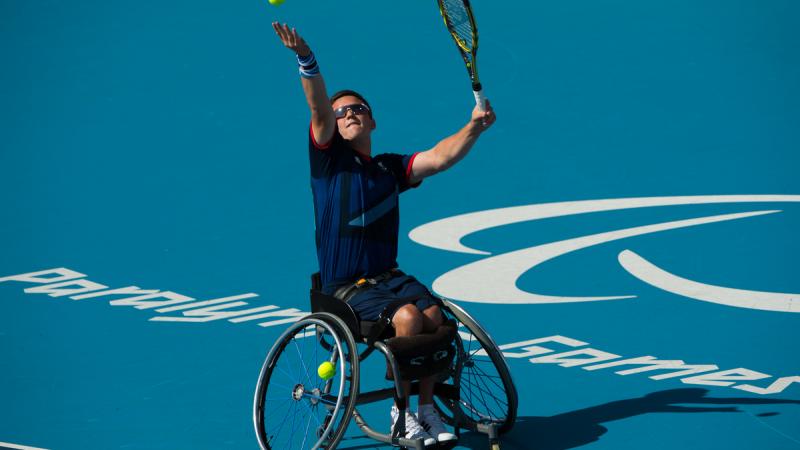 Man in wheelchair serves a ball on a tennis court.