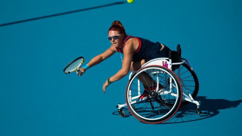 Woman in a wheelchair on a blue tennis court trying to reach a ball