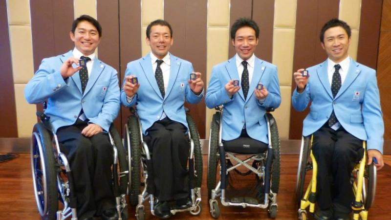 Four men in wheelchairs all wearing blue jackets show pins to the camera, smiling