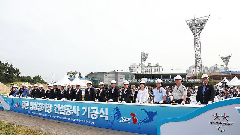 People in suits with construction helmets stand behind a banner in front of a construction area