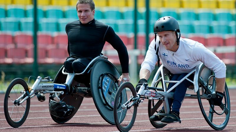 Two male athletes in racing wheelchairs on track, smile to the camera