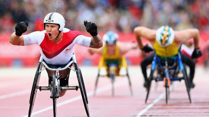 Man in racing wheelchair crosses finish line of a track throwing hands in the air, celebrating