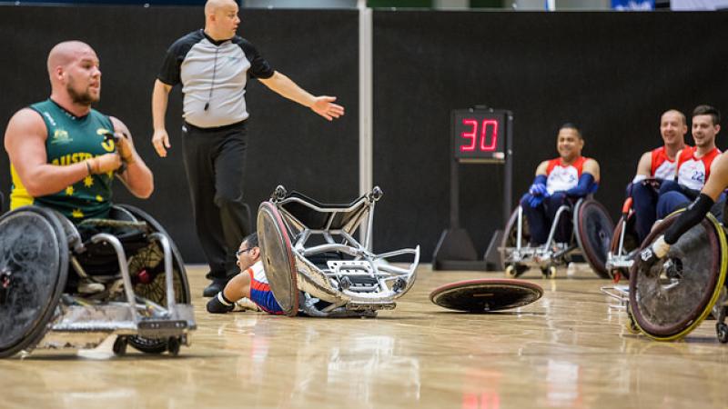 Dressed in green and seated in a wheelchair, a player looks on as another lies face down with his wheel broken from his chair.