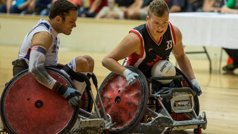 A player wearing blue seated in a wheelchair with a white ball on his lap about to collide with an opponent wearing white.