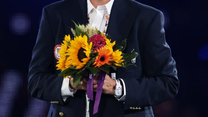 A picture of a smartly dress man, with light trousers and dark jacket holding a bunch of flowers.