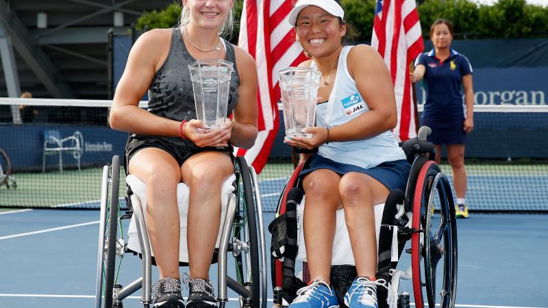 Two players sat in wheelchair holding trophies on their laps. In the background are two USA flags.