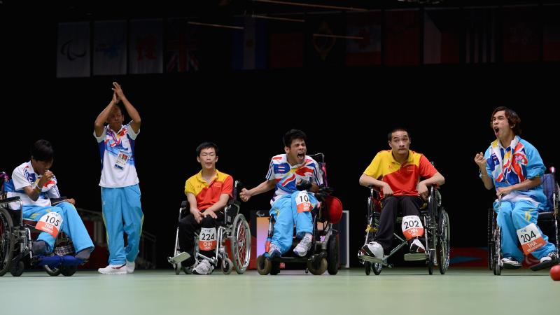 Four men in wheelchairs and one man standing on a field of play with dark background. Three men celebrating
