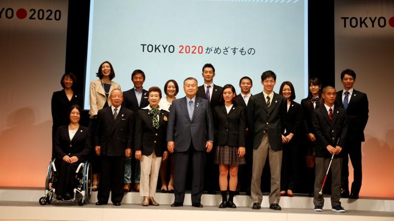 Japanese people standing in a line under a Tokyo 2020 banner