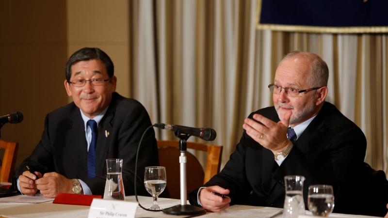 Two men sitting on a podium behind a table with microphones in front of them