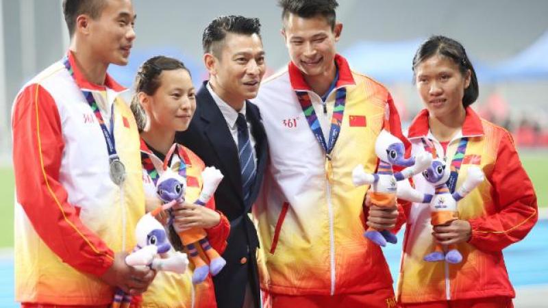 Man in dark suit stands between some athletes on the podium 