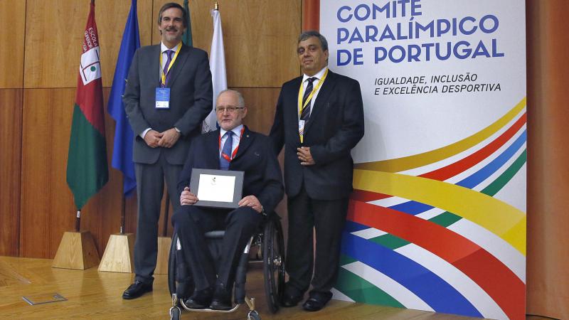 Three men (one in a wheelchair) pose in front of a wooden wall and some flags