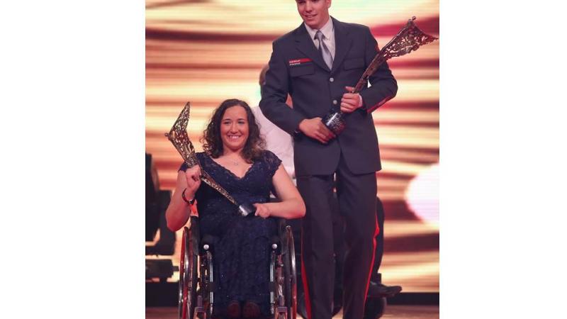 Women in wheelchair, man walking both holding a trophy in their hands