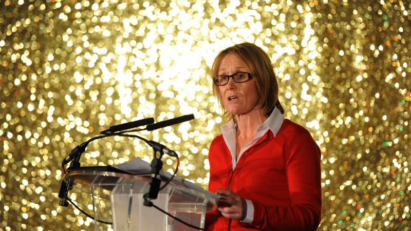 woman in red standing on stage in front of a gold background and addressing the crowd