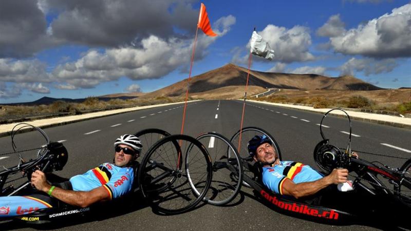Wearing blue lycra outfits, two men lie in their handcycles on an open road with hills in the background.