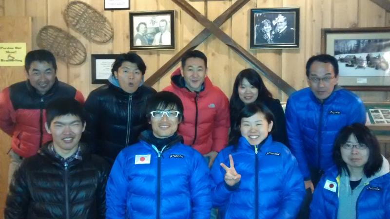 Group shot of six people in front of a wooden wall, smiling to the camera