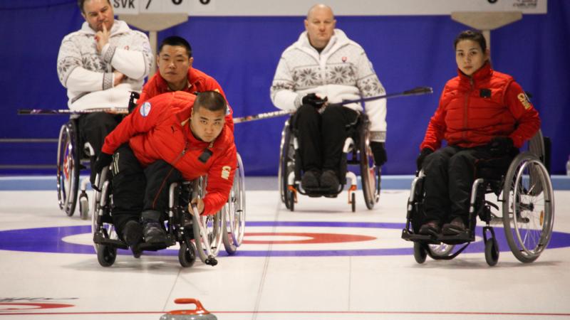 Chinese man in a wheelchair using a stick to throw a curling stone.