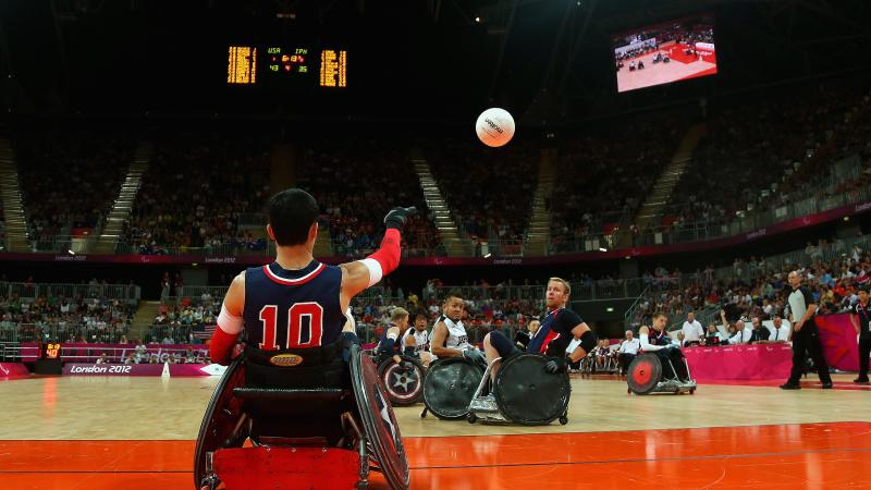 Man in wheelchair throwing a ball, two other men trying to catch it