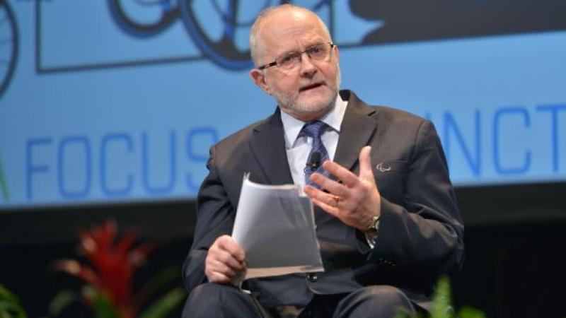 A man seated holding papers in one hand and using his other hand to express his feelings whilst giving a talk.