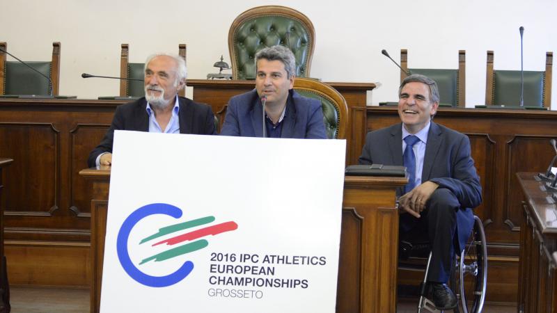 Three men behind a table present the logo for the 2016 IPC Athletics European Championships.