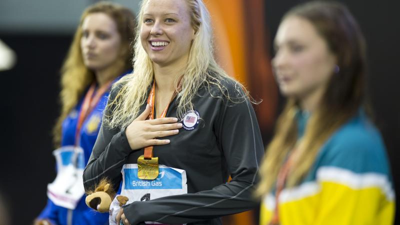 Three women on a podium, one of them smiling, in focus and looking to the camera