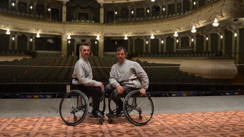 Two men in wheelchairs on a stage in front of empty seats