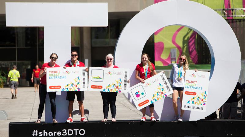 Five persons with big tickets in their hands pose in front of a big sign with the letters TO