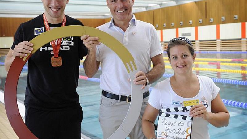 Two men and a woman in a wheelchair pose in front of a swimming pool.