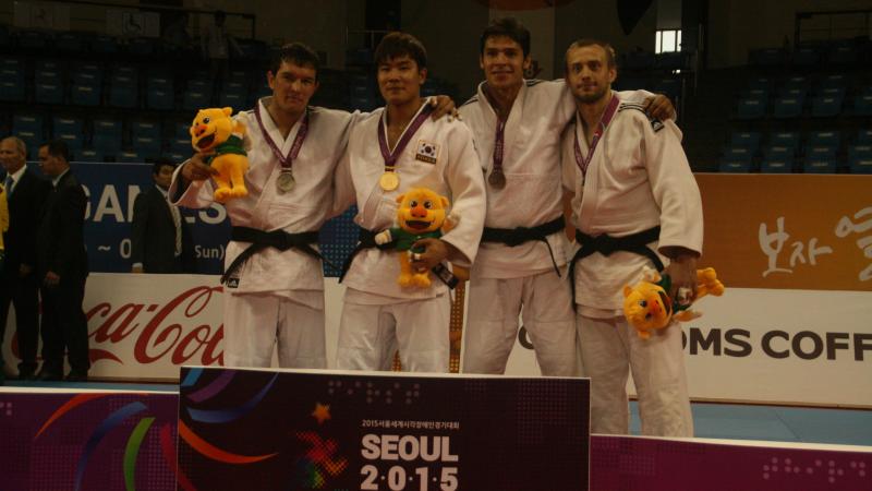 Three men in white training suits stand on a podium with medals