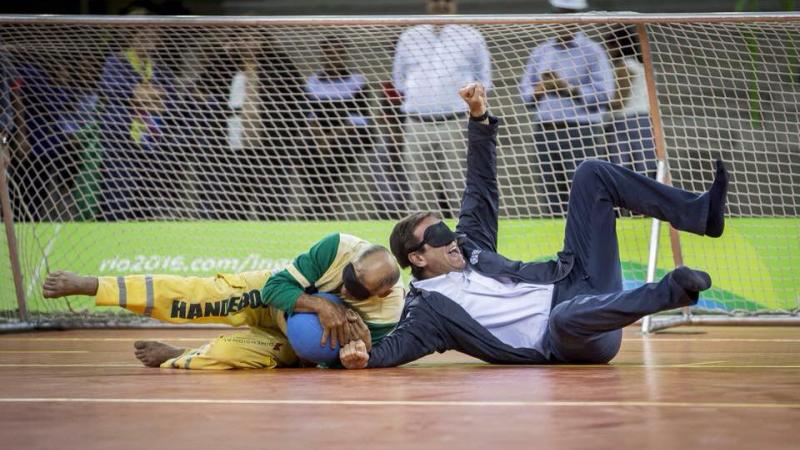 Two blindfolded men, one in a suit lying on a field of play, catching a ball