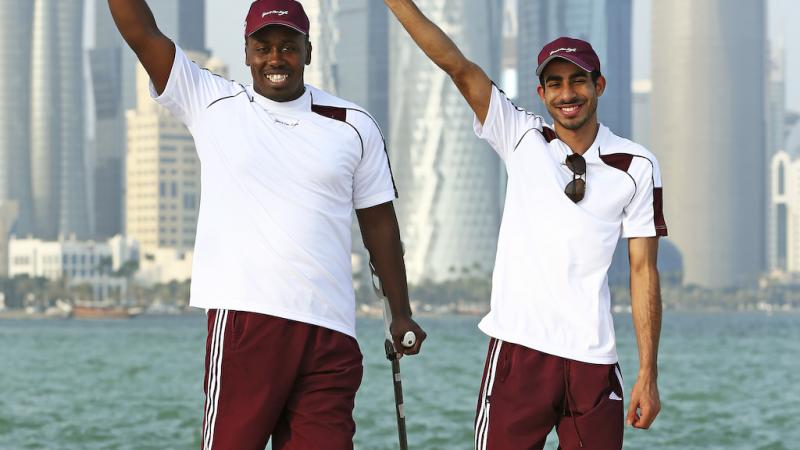 Two men in training suits in front of a skyline, waving