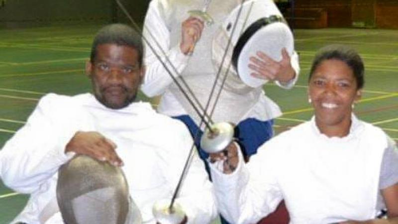Two fencers in a wheelchair holding a sword and an instructor standing in the background. 