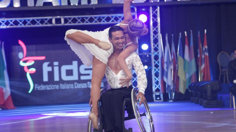 Man in wheelchair holds woman in air while spinning wheelchair.