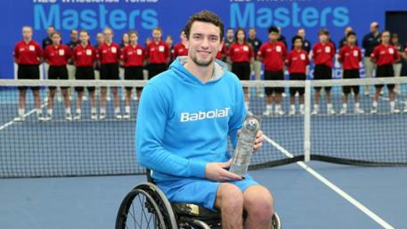 Man in wheelchair on a tennis court, showing a trophy to the camera